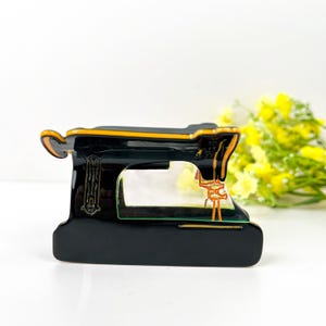 Peut inclure: Figurine décorative de machine à coudre noire et dorée. La figurine a une finition brillante avec des accents orange et verts. La machine à coudre a un design détaillé et est placée sur un fond blanc.