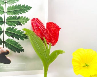 Könnte beinhalten: Ein rotes tulpenförmiges Dekorationsobjekt mit grünen Blättern und Stiel. Die Tulpe hat zwei rote Blütenköpfe. Die Blätter und der Stiel sind hellgrün. Das Objekt befindet sich vor einem weißen Hintergrund mit einem Farn und einer gelben Blume.