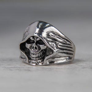 Peut inclure: Bague argentée représentant une tête de mort avec une capuche. Le crâne présente des détails tels que des orbites, une cavité nasale et des dents. La bague a une texture et une esthétique gothique. Un bijou audacieux.