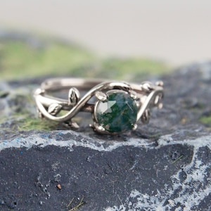 Könnte beinhalten: Ein silberner Ring mit einem grünen Moosachat-Edelstein, der in einem zarten Rankendesign gefasst ist.