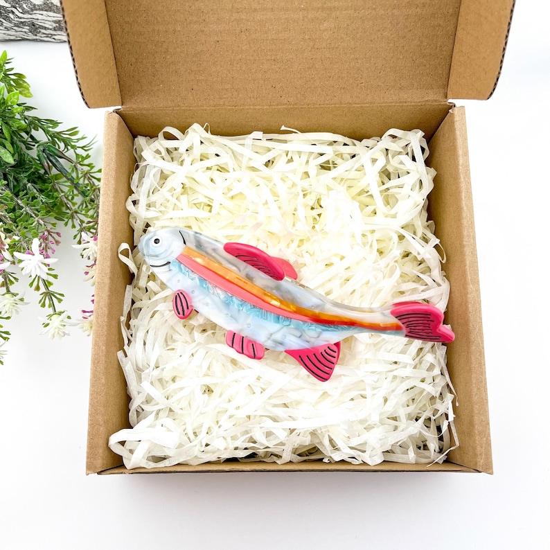 May include: A glass fish figurine with a pink, orange, and white design. The fish is in a cardboard box with white packing peanuts.
