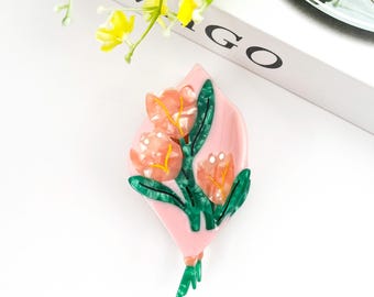 Könnte beinhalten: Eine rosa Blumenbrosche mit drei pfirsichfarbenen Blüten mit gelben Akzenten und grünen Blättern. Die Brosche hat einen rosa Hintergrund und einen grünen Stiel. Die Brosche ist ein einzigartiges Accessoire.