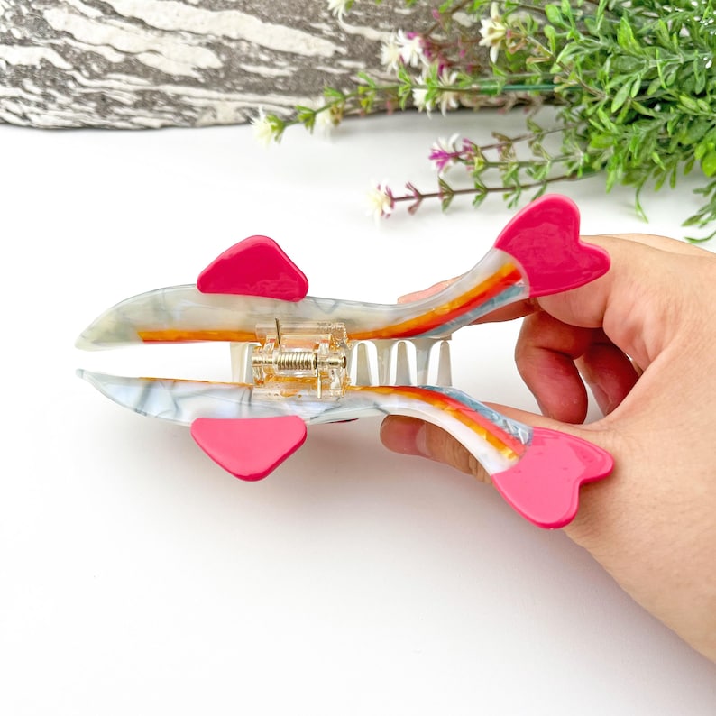 May include: A white and rainbow hair claw with pink heart-shaped ends. The claw has a marble-like pattern and a gold spring.