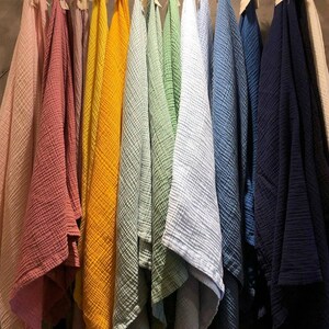 May include: A collection of colorful, textured fabric cloths hanging on a wooden rack. The cloths are in various shades, including pink, coral, yellow, green, blue, and navy. The fabric appears to be a lightweight, crinkled material.