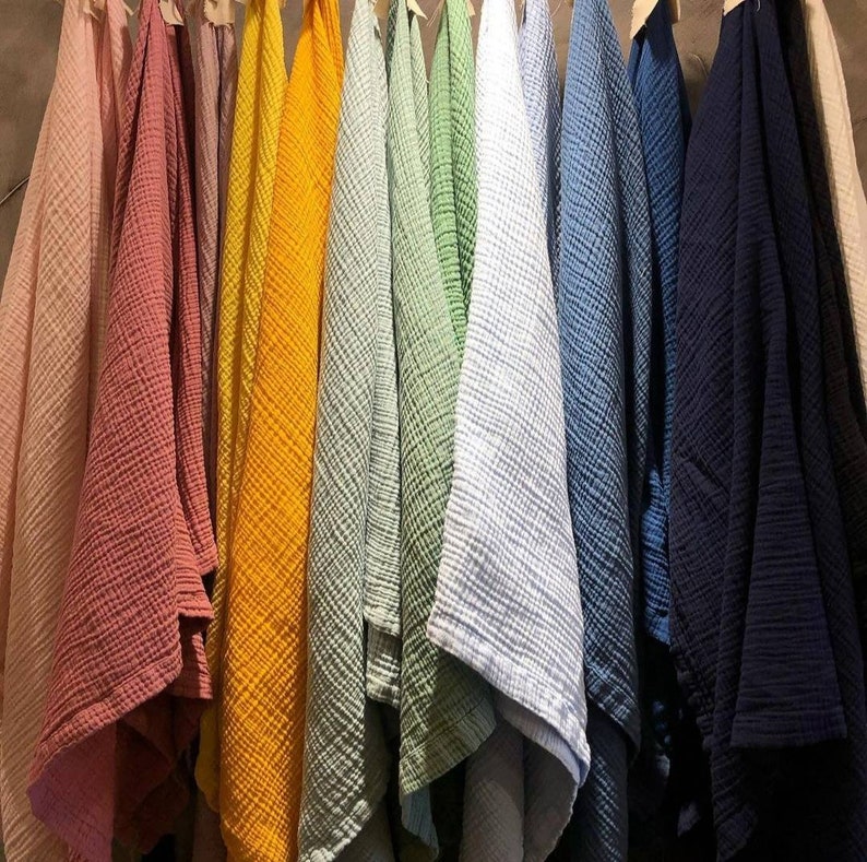 May include: A collection of colorful, textured fabric cloths hanging on a wooden rack. The cloths are in various shades, including pink, coral, yellow, green, blue, and navy. The fabric appears to be a lightweight, crinkled material.