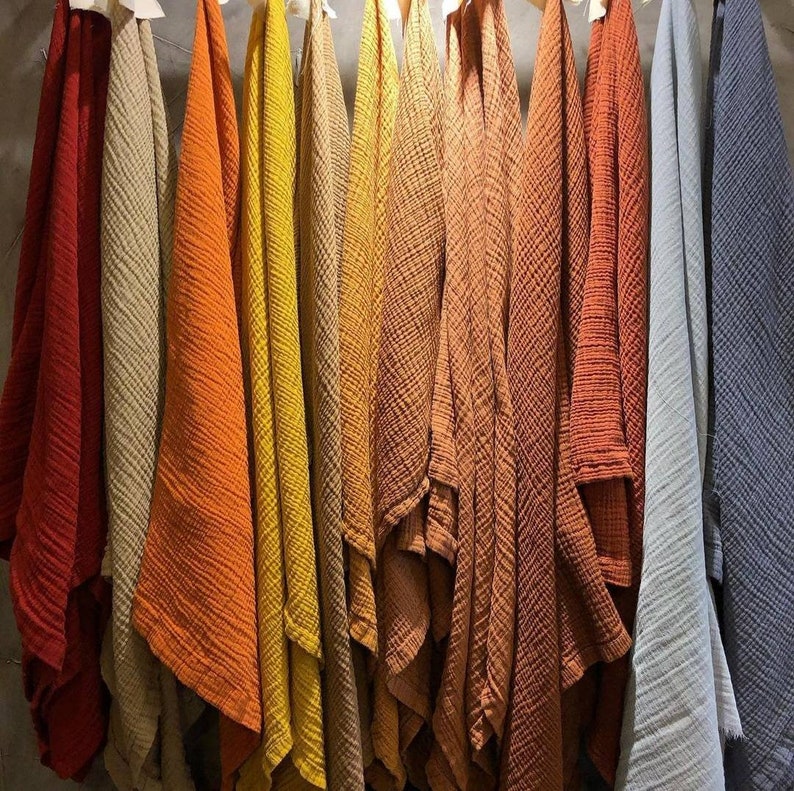 May include: A collection of colorful, textured fabric cloths hanging on wooden hangers. The cloths range in color from deep red to light gray, with shades of orange, yellow, and brown in between. The fabric has a crinkled appearance.