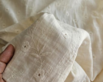 May include: Close-up of off-white fabric with embroidered floral details. The fabric has a delicate texture with small, evenly spaced eyelet cutouts. A hand gently holds a folded section, showcasing the intricate embroidery.