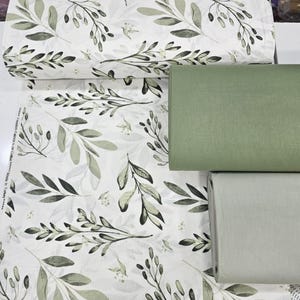 May include: White fabric roll with a botanical leaf pattern in shades of green and gray. Two solid green fabric rolls are also visible. The fabric is likely cotton and suitable for various sewing projects.
