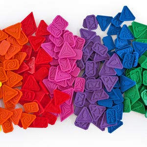 Può includere: Una collezione di colorati pezzi di gioco in un gradiente arcobaleno. I pezzi hanno varie forme, tra cui triangoli e rettangoli, e sono disposti in uno spettro dal giallo al nero. I pezzi hanno disegni in rilievo.