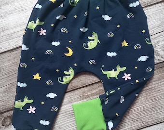 Puede incluir: Pantalones de bebé azules con cintura y puños verdes. Los pantalones tienen un patrón repetido de dragones verdes, estrellas, nubes y arcoíris.