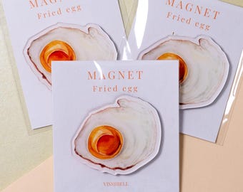 Può includere: Tre calamite raffiguranti un'illustrazione ad acquerello di un uovo fritto. Ogni calamita è montata su una scheda bianca con la scritta "MAGNET Fried egg" in arancione. Le calamite sono confezionate in buste di plastica trasparente.