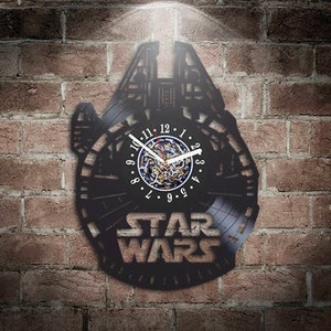 May include: A black vinyl record clock featuring a silhouette of the Millennium Falcon from Star Wars. The clock face is a circular cutout with Roman numeral markings and a working clock mechanism. The words "STAR WARS" are printed in yellow below the clock face.