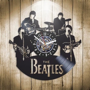 May include: A black vinyl record clock featuring a silhouette of The Beatles band members playing instruments. The clock face is a circular cutout with Roman numeral markings and a clock hand mechanism. The text "The Beatles" is printed below the clock face.