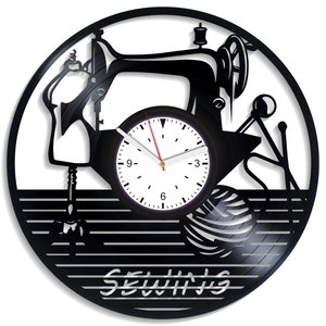 Può includere: Orologio in vinile nero con una silhouette di una macchina da cucire, un rocchetto di filo e tessuto. Il quadrante dell'orologio è bianco con numeri e lancette neri. Le parole "CU­CITURA" sono stampate sotto il quadrante dell'orologio.