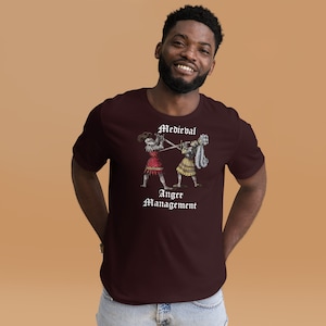 May include: A maroon t-shirt featuring a graphic of two medieval knights fighting with swords. The text "Medieval Anger Management" is printed below the graphic.