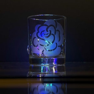 Pode incluir: Um copo de vidro transparente com um desenho de rosa branca fosca. O copo é iluminado por baixo, projetando um brilho azul e roxo.