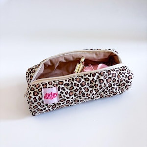 May include: A leopard print makeup bag with a gold lipstick and pink items inside. The bag has a pink label that says "girly bags". The bag is open and has a beige zip.