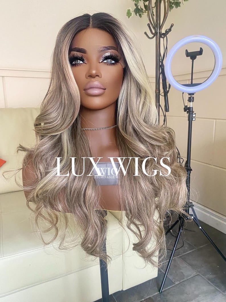 Pu&ograve; includere: Una parrucca lunga e ondulata con radici scure e riflessi biondi &egrave; esposta su una testa di manichino. La parrucca ha un frontale in pizzo ed &egrave; acconciata con ricci sciolti. Il testo "LUX WIGS" &egrave; visibile.