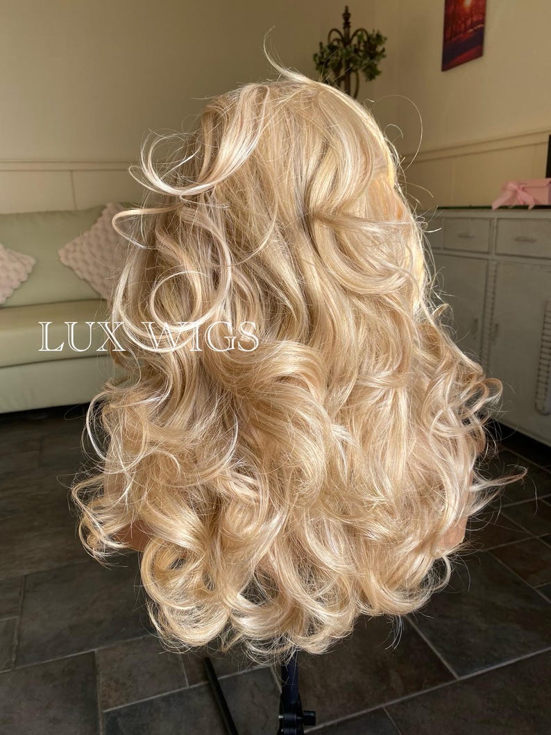 May include: A long, blonde wig with loose curls. The wig is styled in a way that creates volume and movement. The wig is on a mannequin head.