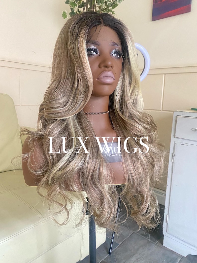 Pu&ograve; includere: Una lunga parrucca ondulata con radici scure e riflessi biondi &egrave; esposta su una testa di manichino. La parrucca ha un frontale in pizzo ed &egrave; acconciata con riccioli sciolti. Il testo "LUX WIGS" &egrave; visibile.