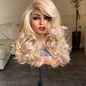 May include: A blonde wig with long, loose curls. The wig is styled with a side part and is worn on a mannequin head. The wig is on a black stand. The text "LUX WIGS" is visible on the wig.