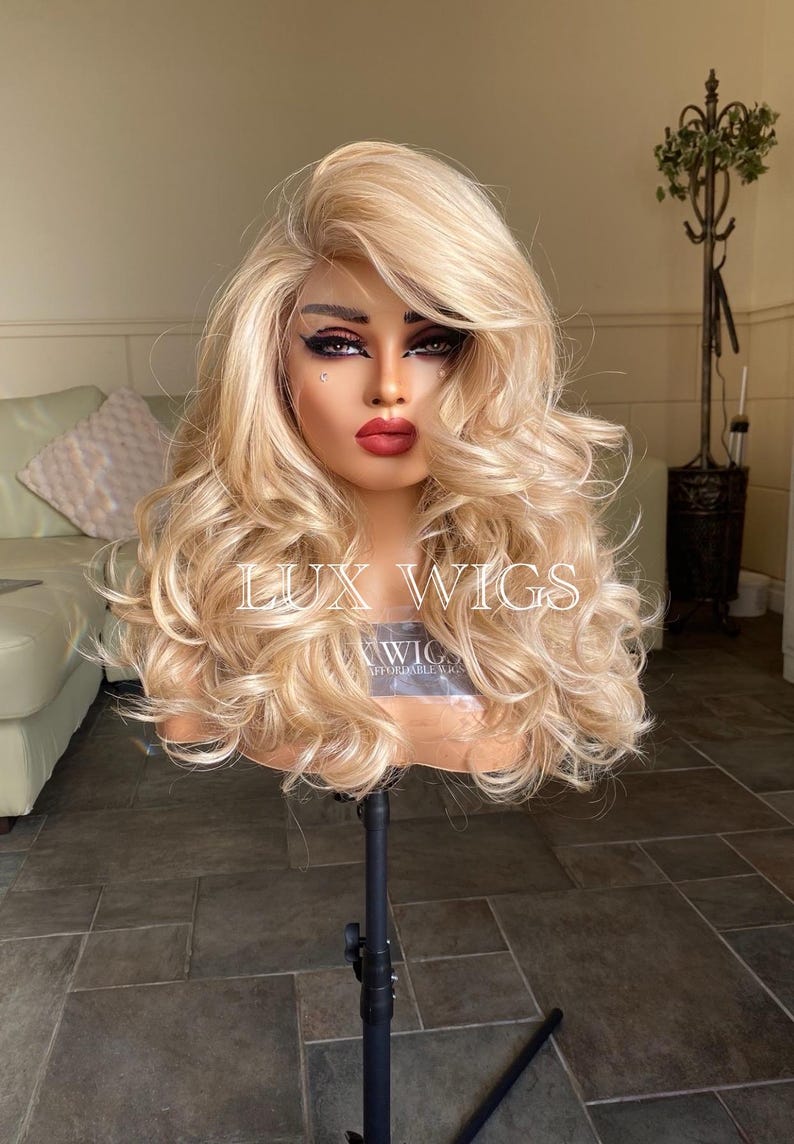 May include: A blonde wig with long, loose curls. The wig is styled with a side part and is worn on a mannequin head. The wig is on a black stand. The text "LUX WIGS" is visible on the wig.