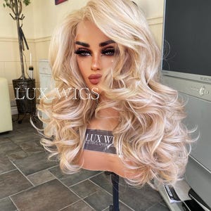 May include: A blonde wig with loose curls and a light brown base. The wig is on a stand and has a label that says "Lux Wigs Beautiful Affordable Wigs".