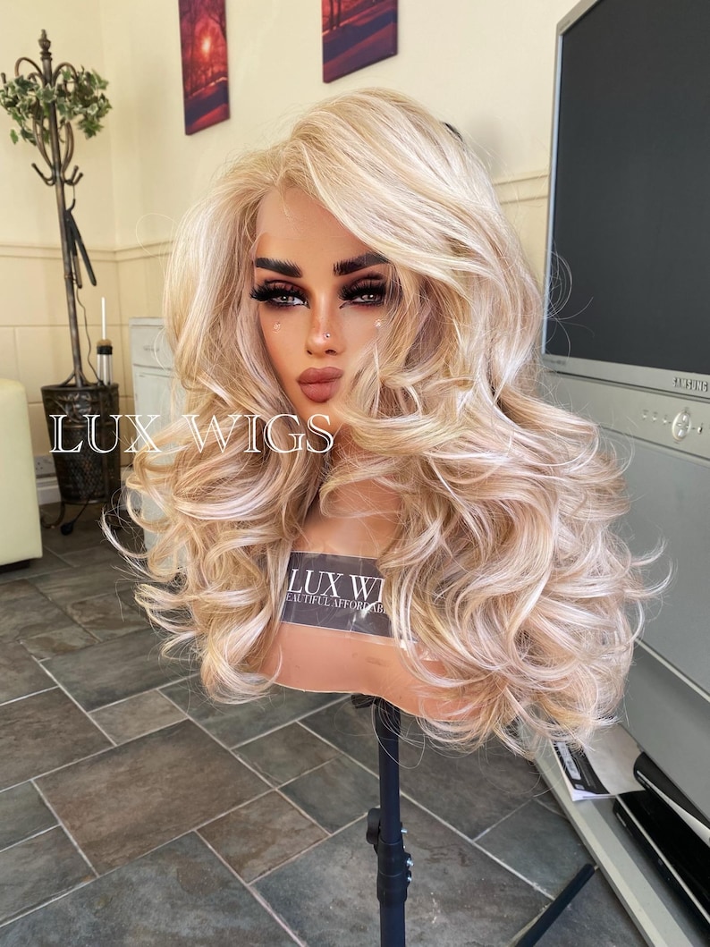 May include: A blonde wig with loose curls and a light brown base. The wig is on a stand and has a label that says "Lux Wigs Beautiful Affordable Wigs".
