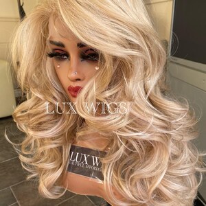 May include: A long, wavy, blonde wig with a light brown base. The wig is styled with loose curls and is worn on a mannequin head. The wig is labeled "LUX WIGS" and "BEAUTIFUL AFFORDABLE WIGS".