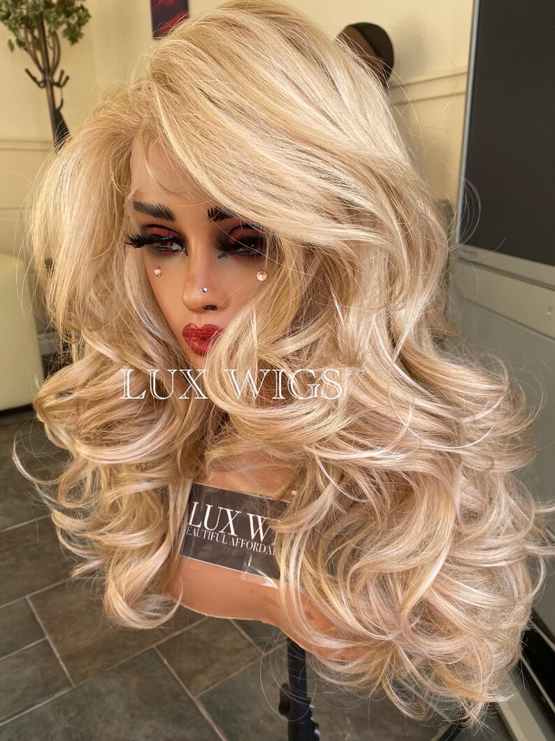 May include: A long, wavy, blonde wig with a light brown base. The wig is styled with loose curls and is worn on a mannequin head. The wig is labeled "LUX WIGS" and "BEAUTIFUL AFFORDABLE WIGS".