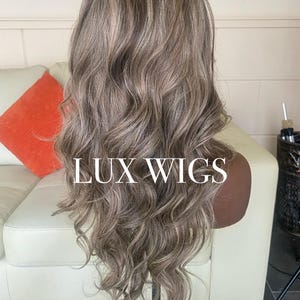 Pu&ograve; includere: Una parrucca lunga e ondulata con una miscela di riflessi grigi e marrone chiaro. La parrucca &egrave; acconciata su una testa di manichino, con il testo "LUX WIGS" visualizzato. I capelli hanno un aspetto naturale e fluente.