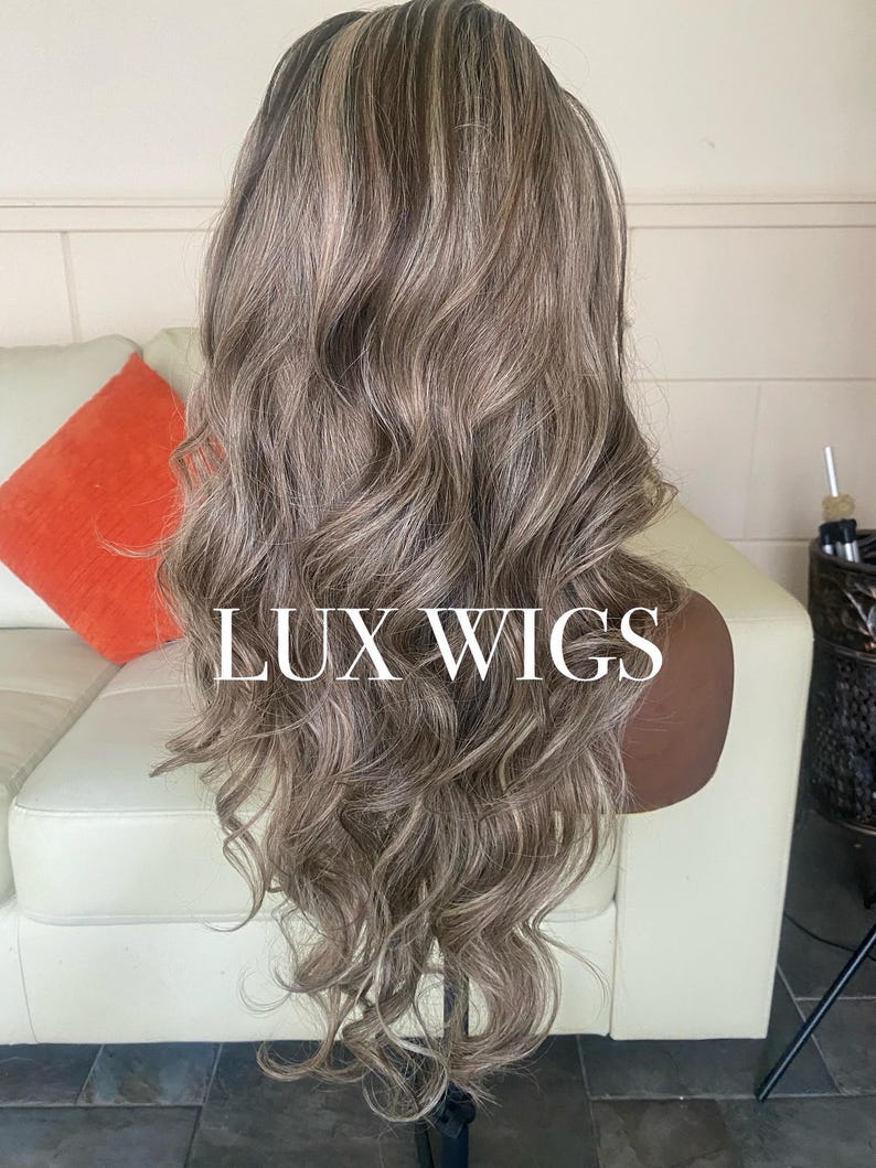Pu&ograve; includere: Una parrucca lunga e ondulata con una miscela di riflessi grigi e marrone chiaro. La parrucca &egrave; acconciata su una testa di manichino, con il testo "LUX WIGS" visualizzato. I capelli hanno un aspetto naturale e fluente.
