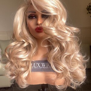 May include: A blonde wig with loose curls on a wig stand. The wig is styled with a side part and has a natural, flowing look. The wig is labeled "LUX WIG" and "Beautiful Affordable Wigs".