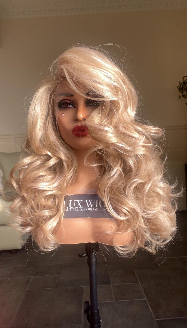 May include: A blonde wig with loose curls on a wig stand. The wig is styled with a side part and has a natural, flowing look. The wig is labeled "LUX WIG" and "Beautiful Affordable Wigs".