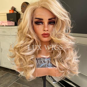 May include: A blonde wig with loose curls on a wig stand. The wig is styled with a side part and has a light blonde color. The wig stand is black and has a round base. The wig is labeled "LUX WIGS" and "LUX WIG BEAUTIFUL AFFORDABLE".