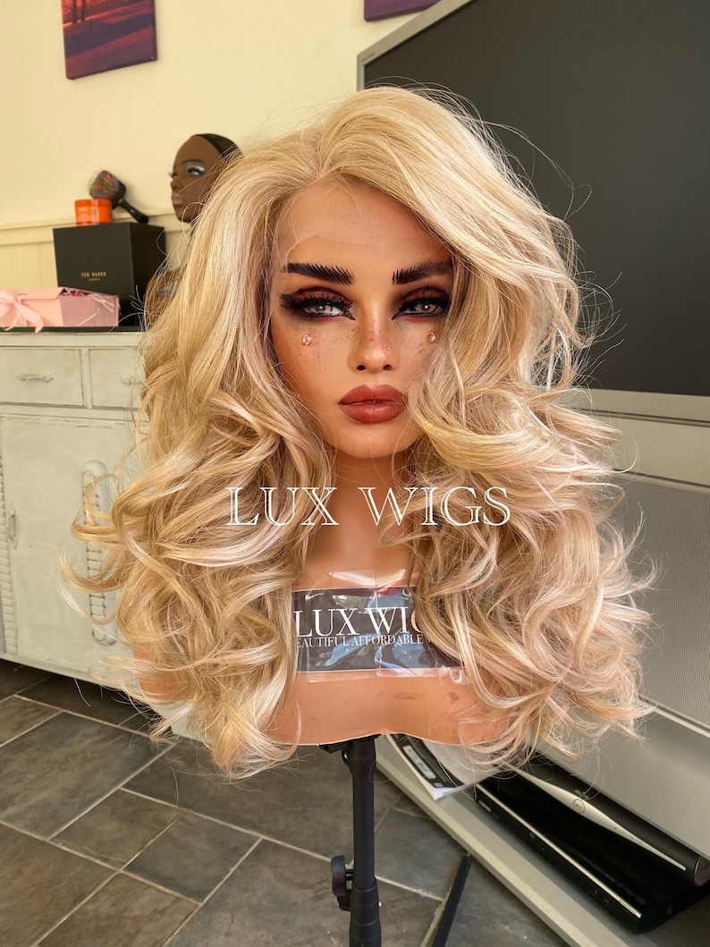 May include: A blonde wig with loose curls on a wig stand. The wig is styled with a side part and has a light blonde color. The wig stand is black and has a round base. The wig is labeled "LUX WIGS" and "LUX WIG BEAUTIFUL AFFORDABLE".