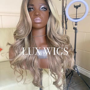 Pu&ograve; includere: Una lunga parrucca ondulata con radici scure e riflessi biondi &egrave; esposta su una testa di manichino. La parrucca &egrave; acconciata con riccioli sciolti e presenta il testo "LUX WIGS" sulla parte anteriore. Un anello luminoso &egrave; posizionato a destra.