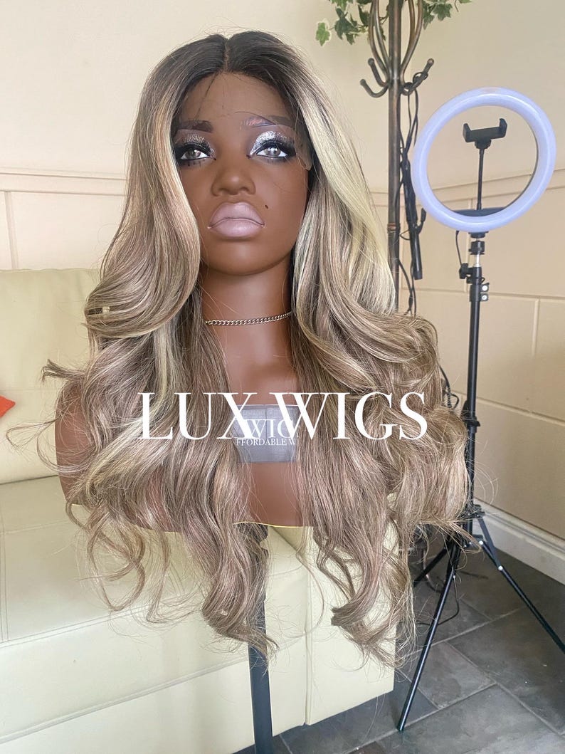 Pu&ograve; includere: Una lunga parrucca ondulata con radici scure e riflessi biondi &egrave; esposta su una testa di manichino. La parrucca &egrave; acconciata con riccioli sciolti e presenta il testo "LUX WIGS" sulla parte anteriore. Un anello luminoso &egrave; posizionato a destra.