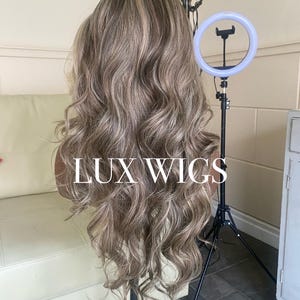 Pu&ograve; includere: Una lunga parrucca ondulata con riflessi biondi e castano chiaro. La parrucca &egrave; esposta su una testa di manichino, con il testo "LUX WIGS". Un anello luminoso e un treppiede sono visibili sullo sfondo.