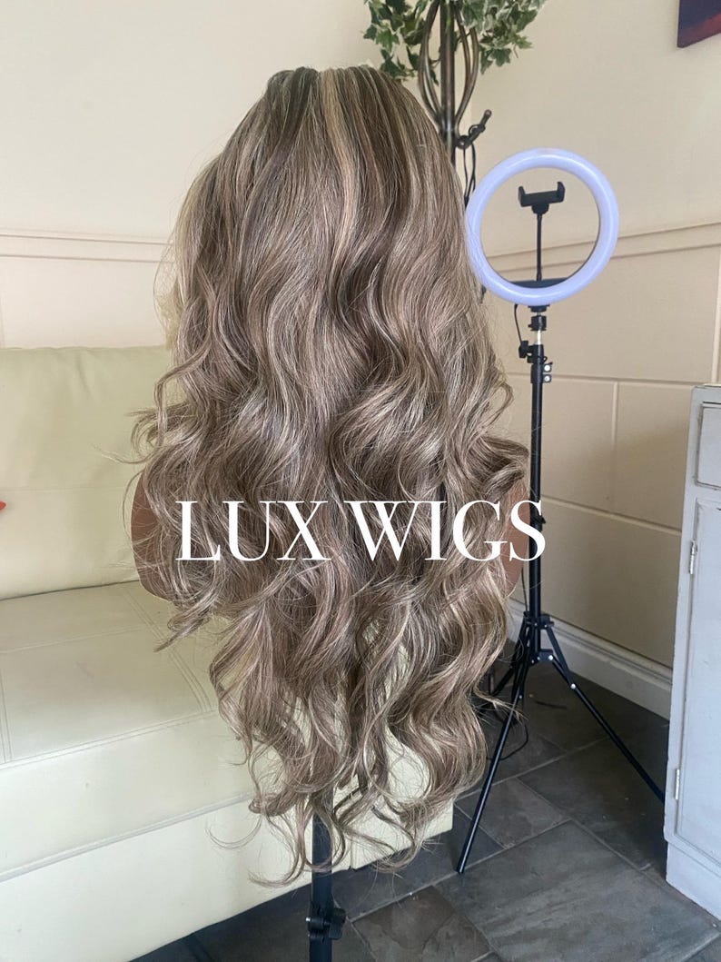 Pu&ograve; includere: Una lunga parrucca ondulata con riflessi biondi e castano chiaro. La parrucca &egrave; esposta su una testa di manichino, con il testo "LUX WIGS". Un anello luminoso e un treppiede sono visibili sullo sfondo.