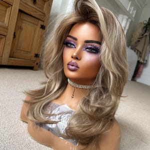 May include: A mannequin head featuring shoulder-length, layered blonde hair with darker roots. The mannequin has stylized makeup with purple glitter eyeshadow, defined eyebrows, and mauve lipstick, and wears a silver rhinestone choker necklace.