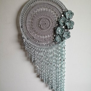 May include: A gray and blue crocheted dreamcatcher with a beaded fringe and crocheted flowers.