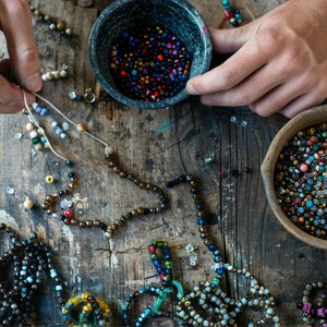 May include: A collection of small, multicolored glass beads in two round bowls and several partially assembled strands of beads. The beads feature various colors including blue, red, yellow, and black, arranged in loose patterns on a wooden surface.