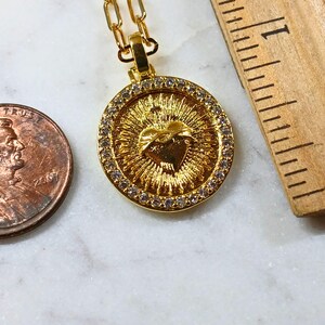 May include: A gold-toned circular pendant necklace featuring a central Sacred Heart emblem with radiating sunburst lines, surrounded by a border of small clear stones. A US penny and a wooden ruler showing 1 inch are placed next to the pendant.
