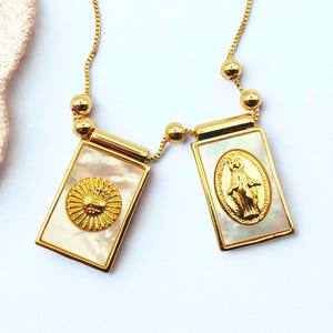 Puede incluir: Collar dorado con dos colgantes rectangulares. Cada colgante tiene una incrustación de nácar. Uno presenta un diseño de sol, el otro una figura religiosa. El collar tiene una cadena fina y cuentas doradas.