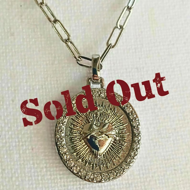 May include: A silver-toned necklace featuring a circular pendant with a central embossed heart surrounded by radiating sunburst lines and a border of small clear stones. The text Sold Out is overlaid in red across the center of the pendant.