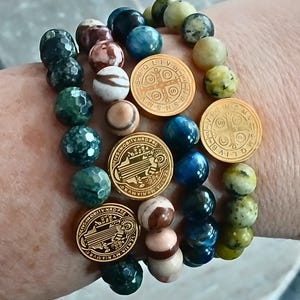 May include: Three beaded bracelets with gold-colored medallions. The bracelets feature a variety of stone beads in shades of green, brown, blue, and cream. The medallions have embossed text and designs. The bracelets are worn on a wrist.