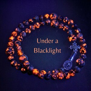 May include: A circular rosary made of dark beads that glow with bright orange and yellow spots under ultraviolet light. The rosary features a silver-toned metal crucifix and a circular medal. The text Under a Blacklight is centered.