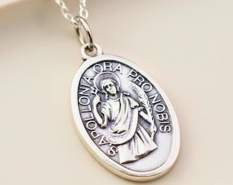 May include: A silver-toned oval pendant necklace featuring a relief image of a figure holding a palm frond and a pincer tool. The text "SANCTA APOLLONIA ORA PRO NOBIS" is embossed around the central figure on the pendant, which hangs from a delicate chain.