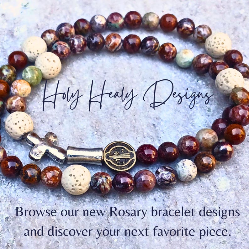 May include: A beaded bracelet featuring brown, red, and cream-colored stone beads, interspersed with porous white beads. It includes a silver-toned cross charm and a circular medal. Text reads: Holy Healy Designs. Browse our new Rosary bracelet designs and discover your next favorite piece.
